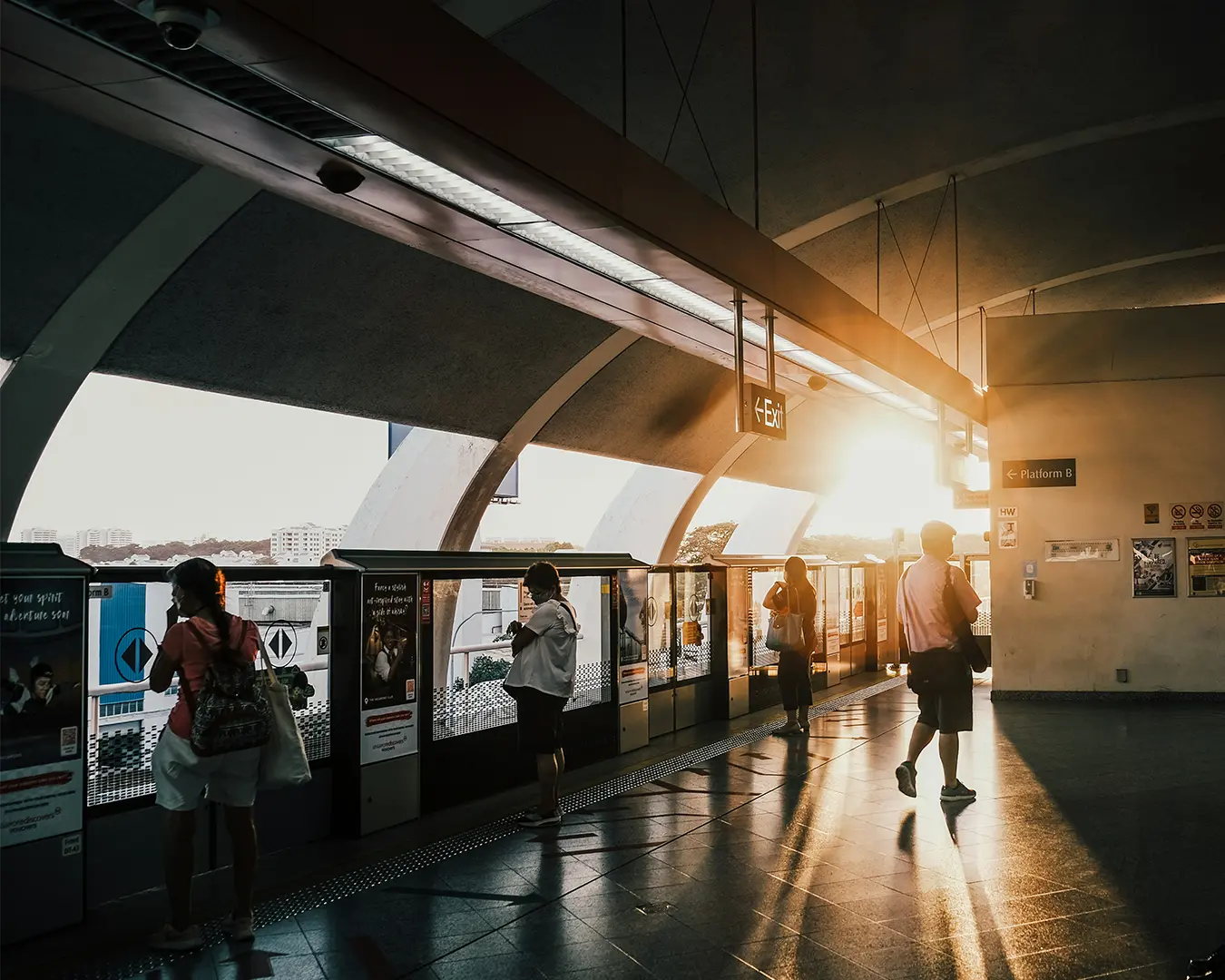 A group of commuters standing on a train platform, patiently waiting for the next train to arrive. Some are looking at their phones while others glance down the tracks, capturing the everyday rhythm of urban public transport.