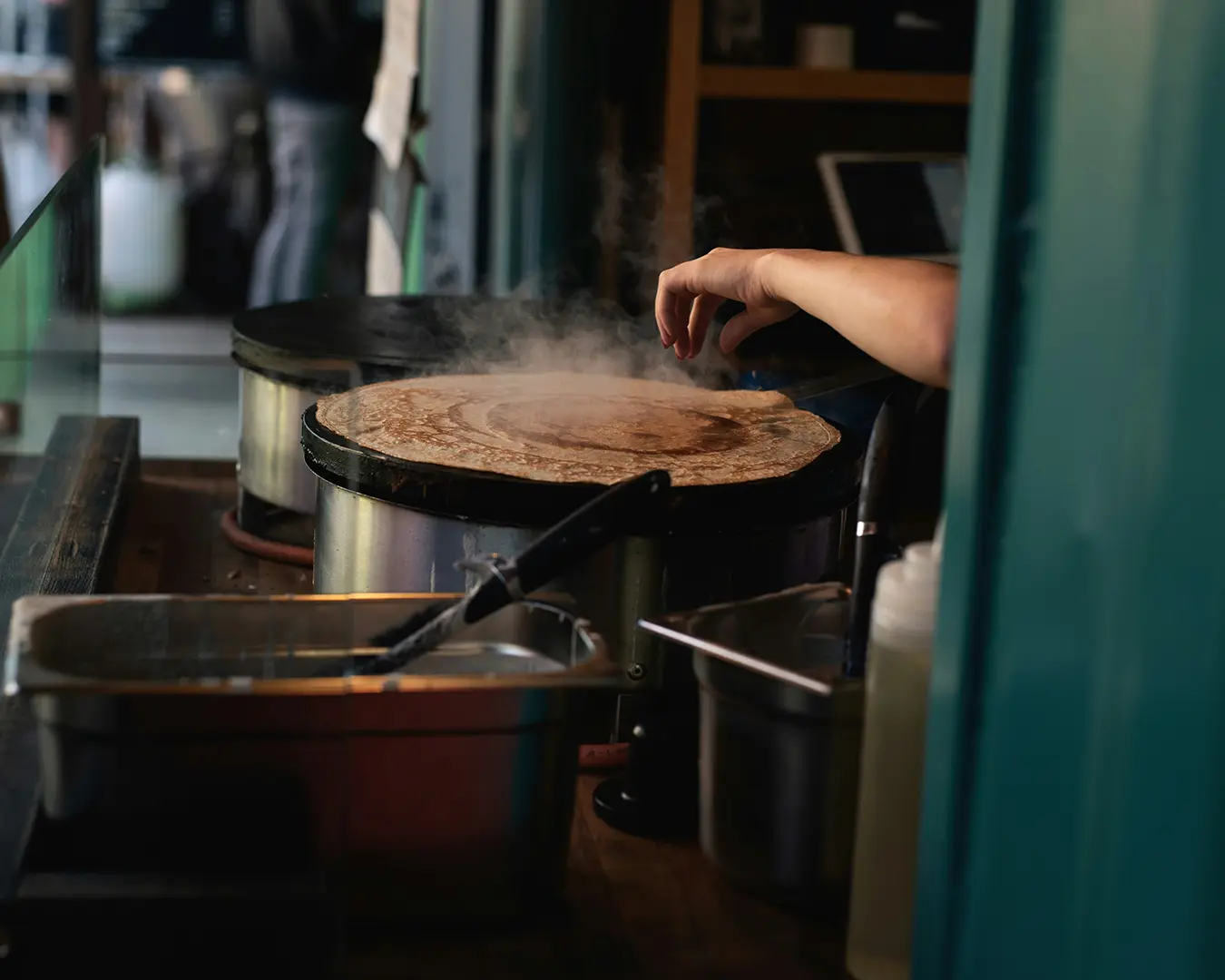 A close-up of a cook’s hand skillfully spreading batter on a hot griddle, preparing crispy tosei under warm night lighting, with steam rising in a bustling street food setting.