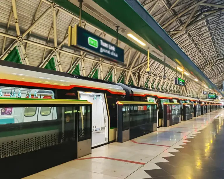 A clean and well-lit MRT train station platform with commuters waiting in an orderly line, train tracks running alongside, and digital signboards displaying arrival times.