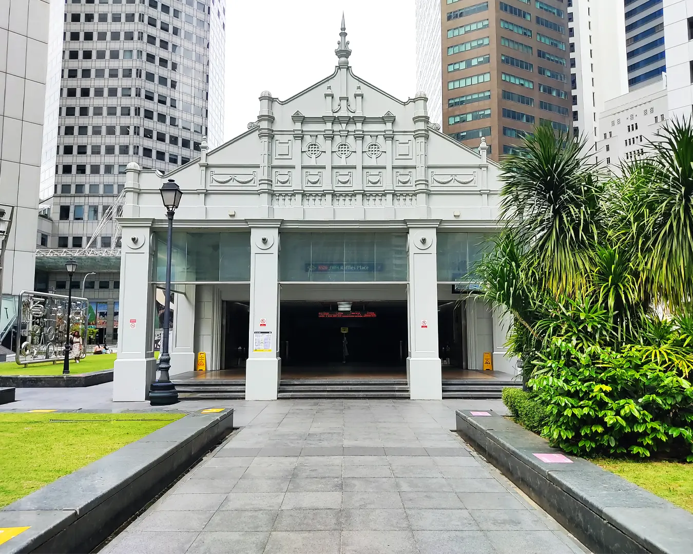 The front entrance area of Raffles Place MRT station surrounded by tall modern office buildings in Singapore’s central business district during the day.