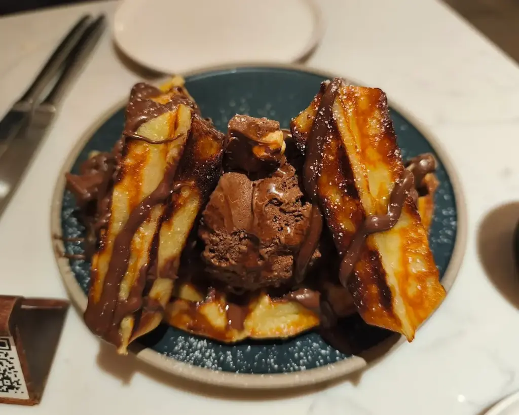 A freshly made chocolate waffle topped with a generous scoop of creamy ice cream, slowly melting into the warm waffle. Rich chocolate drizzle cascades over the edges, creating an indulgent and inviting dessert scene