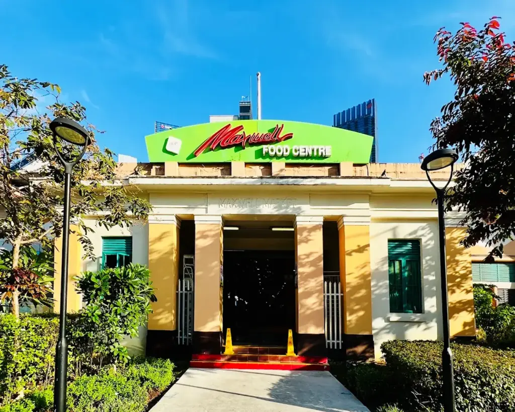 The bustling exterior of Maxwell Food Centre, with people walking in and out of the hawker centre. The scene reflects a lively local atmosphere, with signage visible and the surrounding area filled with movement and anticipation of good food.