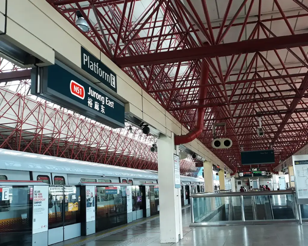 Platform E at Jurong East MRT station with a train approaching, passengers waiting along the platform in a clean and well-lit urban transit environment.
