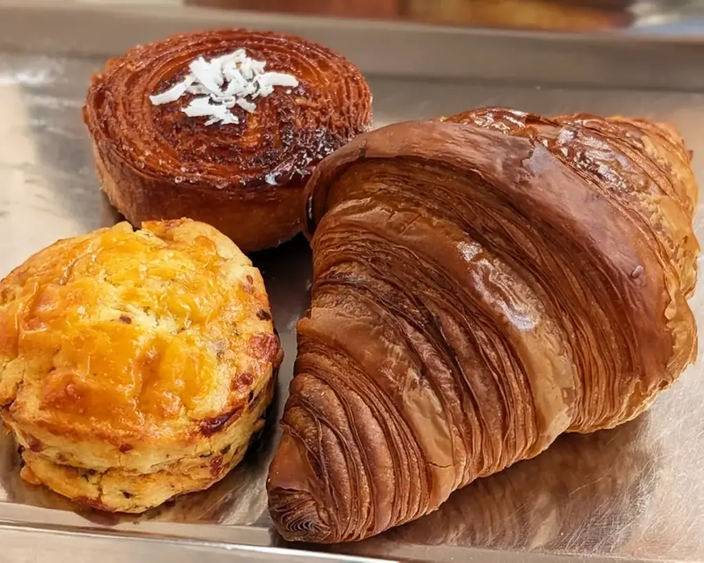 A golden-brown croissant-muffin with flaky, layered edges and a soft, buttery interior. Fresh out of the oven, its crisp outer texture contrasts with the light and airy centre, giving off a warm, comforting bakery feel.