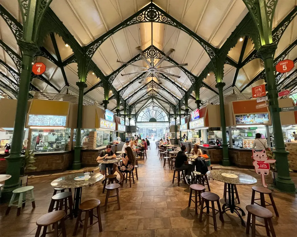 A bustling Singapore food centre filled with diners seated at communal tables, enjoying different hawker dishes while vendors prepare food at surrounding stalls.