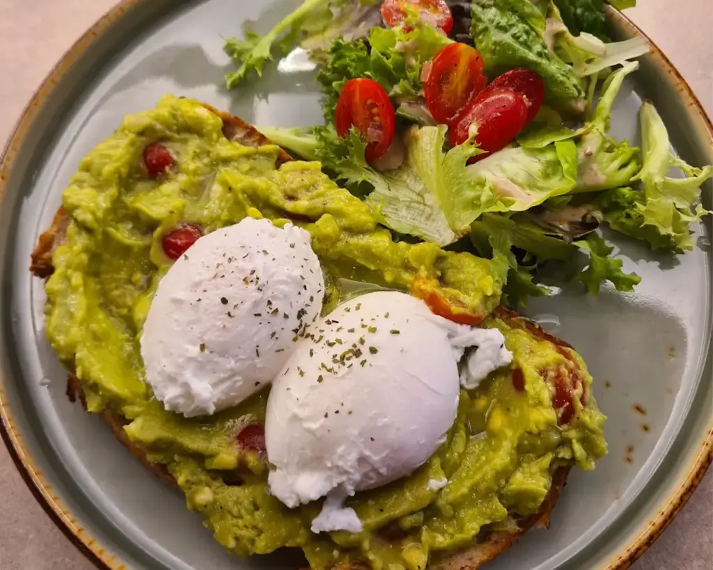 A slice of toasted sourdough bread topped with creamy mashed avocado, spread evenly and garnished with light seasoning. The crispy base and smooth topping create a simple yet wholesome brunch dish.