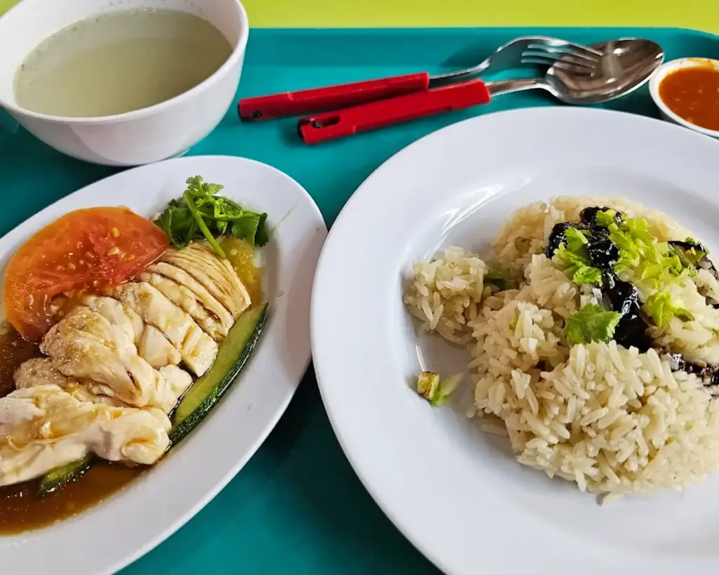 A neatly plated serving of chicken rice placed on a dining table, featuring tender poached chicken, fragrant rice, and small bowls of chilli sauce and ginger paste.