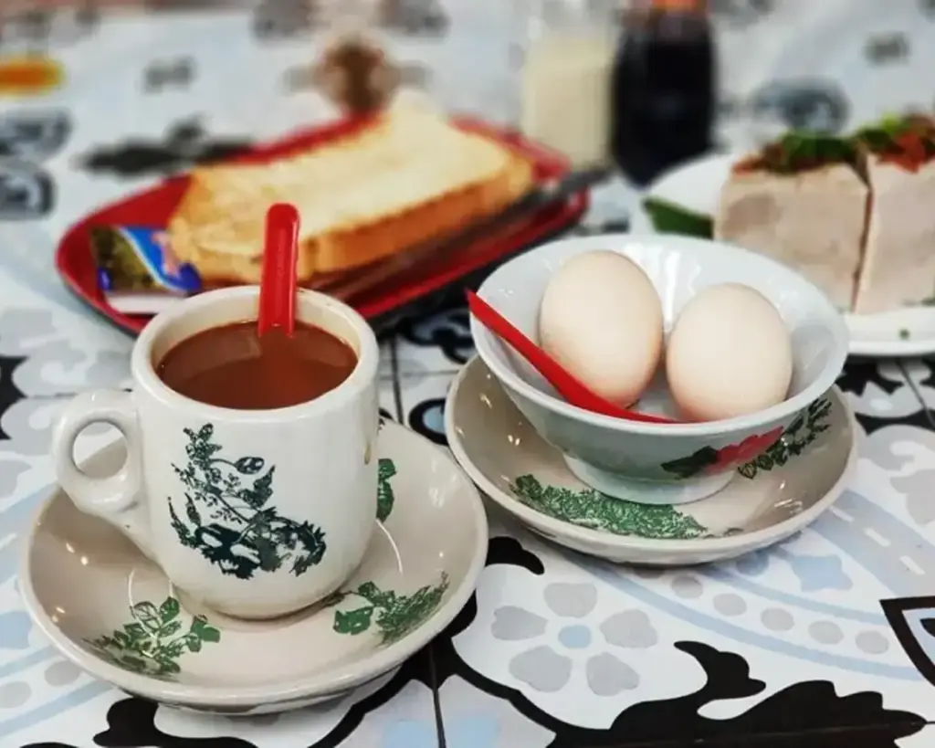 A simple traditional breakfast setting with a warm cup of coffee placed beside a bowl containing two soft-boiled eggs, often served with soy sauce and pepper.