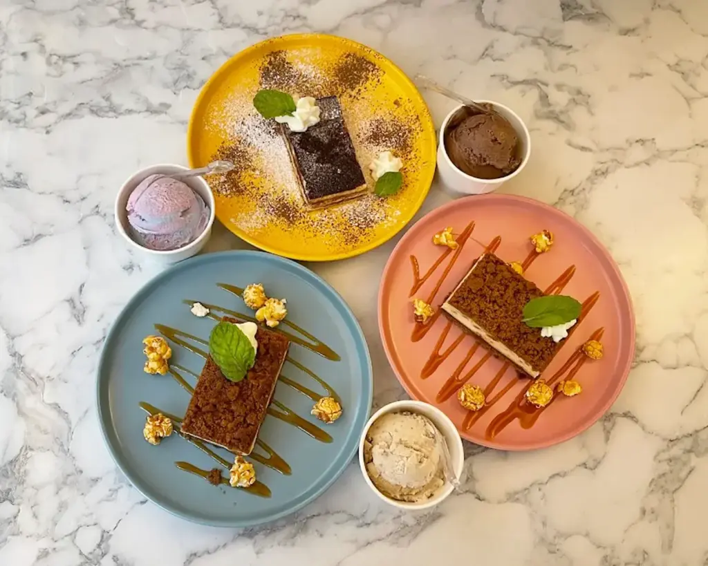 An assortment of three different cake slices arranged on a table, each with distinct textures and colours, paired with a scoop of ice cream. The setting feels cosy and indulgent, perfect for sharing or a dessert treat.