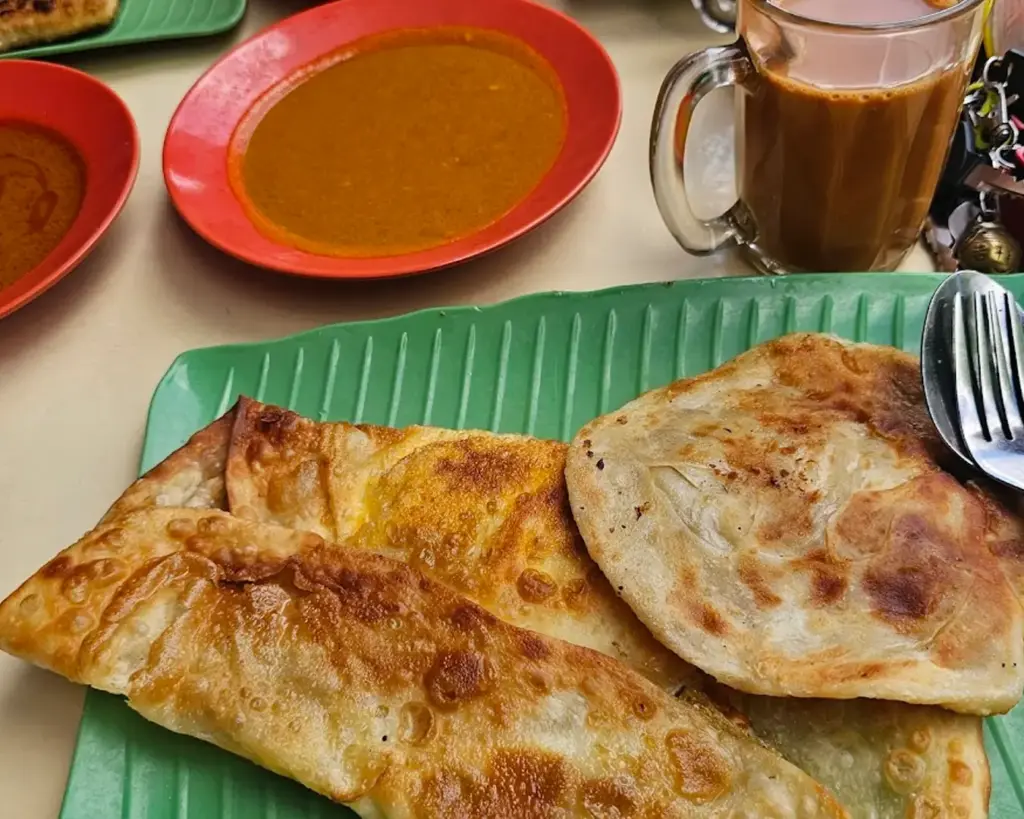 Flaky roti prata served on a plate with a side of curry, paired with a frothy glass of teh tarik, capturing a classic breakfast or supper scene.