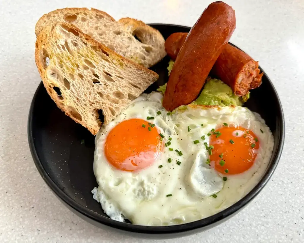 A plated brunch dish featuring eggs benedict with a perfectly cooked sous vide egg resting on a base, accompanied by smoked sausage on the side. The dish is neatly presented, combining rich textures and a satisfying balance of flavours.