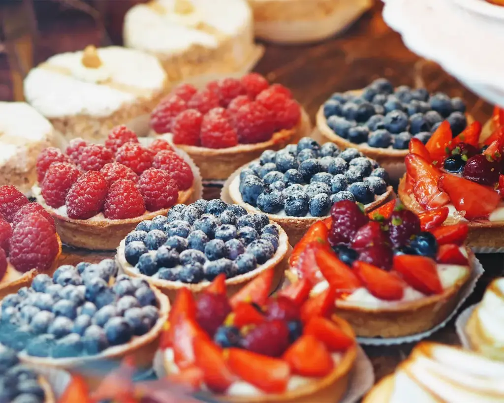 Colourful assortment of fruit tarts with glossy fresh fruits such as strawberries, kiwi, and blueberries arranged on buttery pastry shells filled with smooth custard.