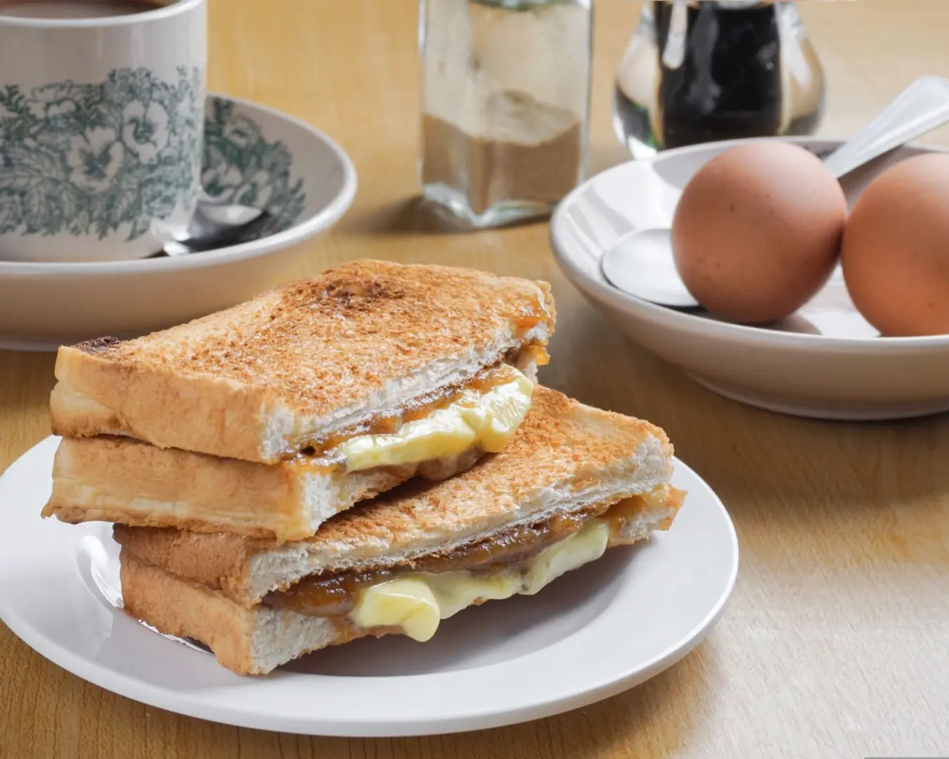 Toasted bread with melted butter and kaya on a plate, beside soft-boiled eggs, a cup of tea, and a sugar jar on a wooden table. Cozy breakfast scene.