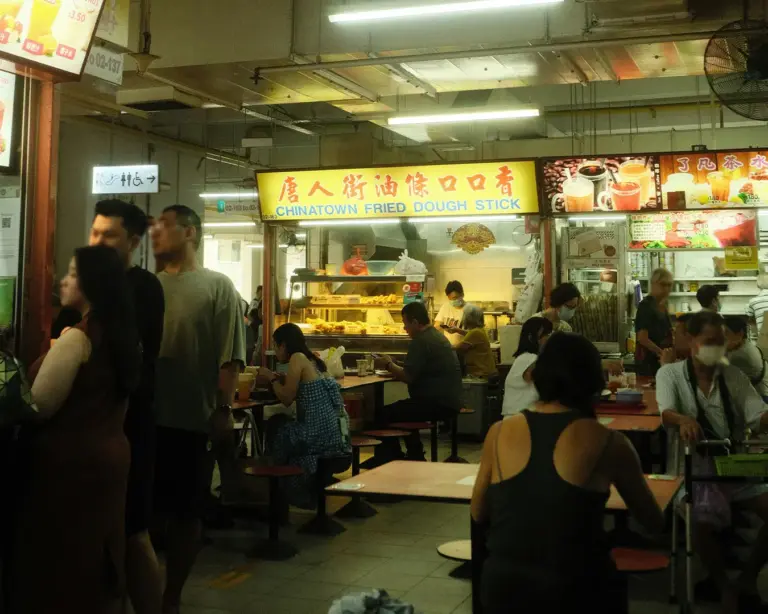 A bustling hawker center with a brightly lit food stall sign reading "Chinatown Fried Dough Stick." Patrons sit and queue, creating a lively atmosphere.