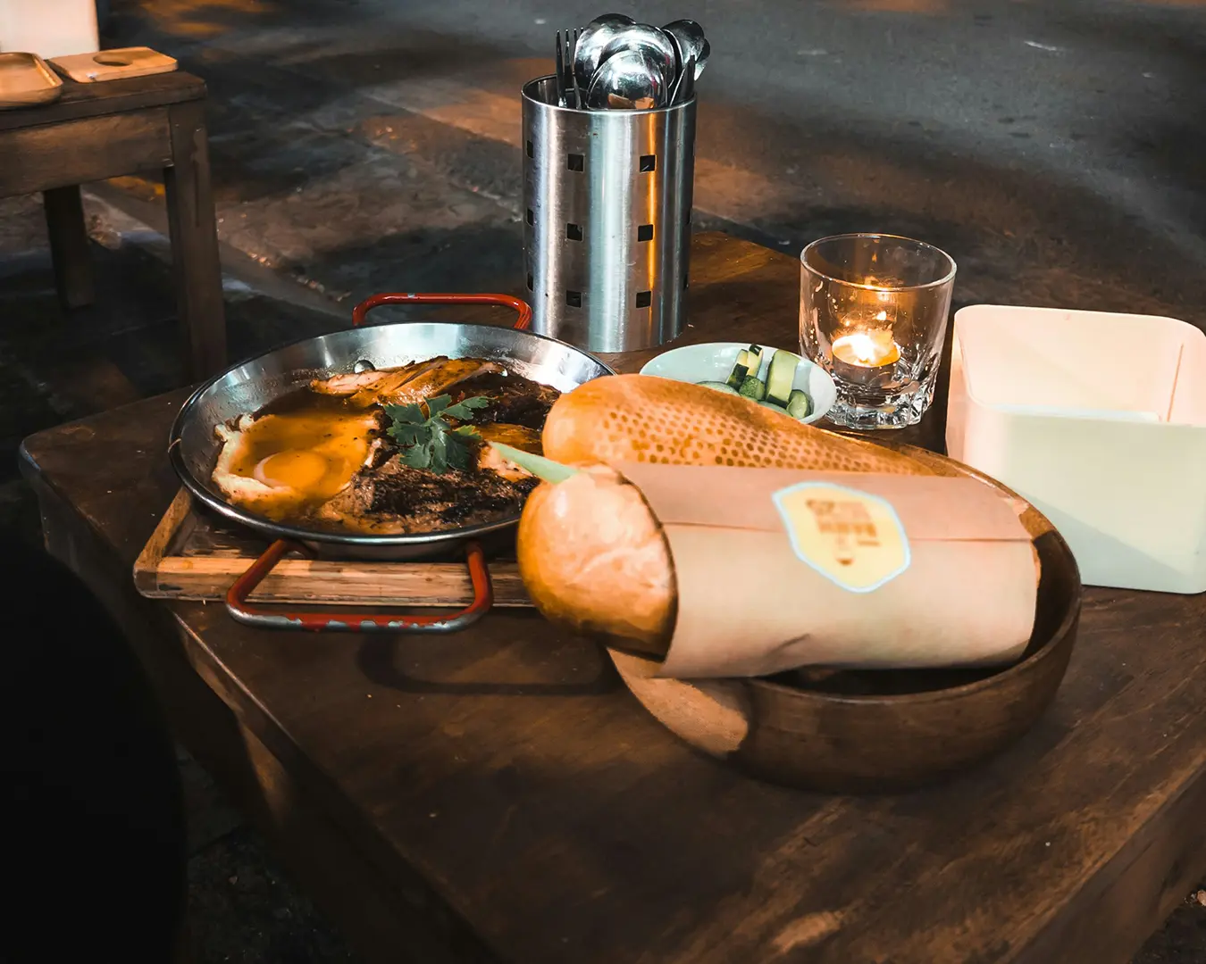 A cozy setting with a wooden table holding a fish dish, baguette wrapped in paper, metal utensil holder, and a lit candle, conveying warmth.