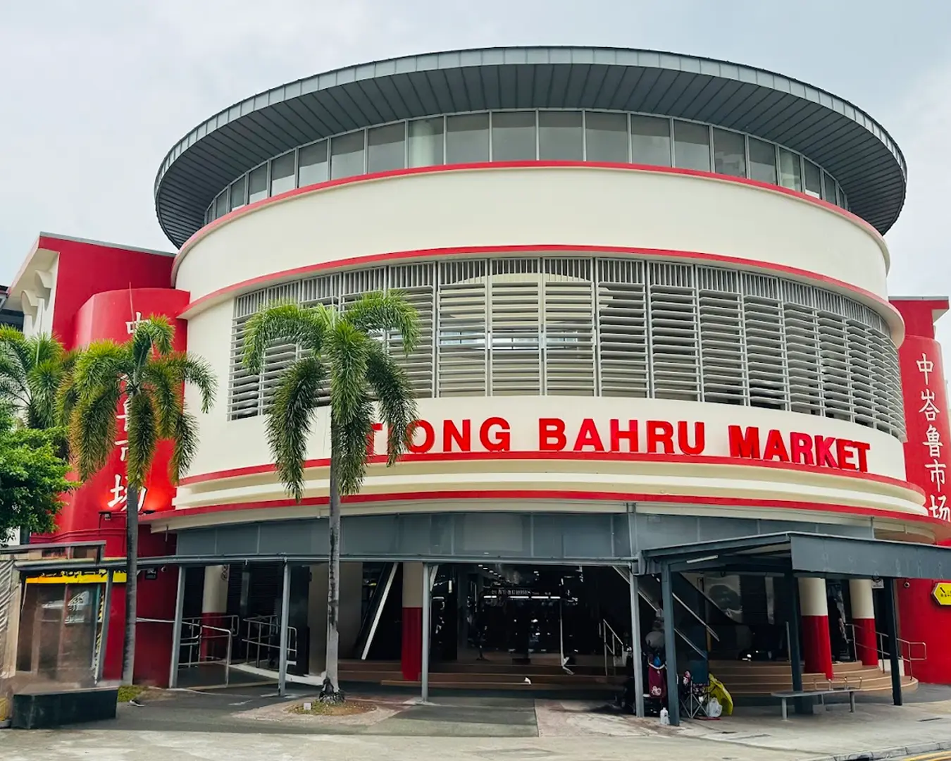 Front view of Tiong Bahru Market, a circular building with red accents and shuttered windows, flanked by palm trees, conveying a vibrant atmosphere.