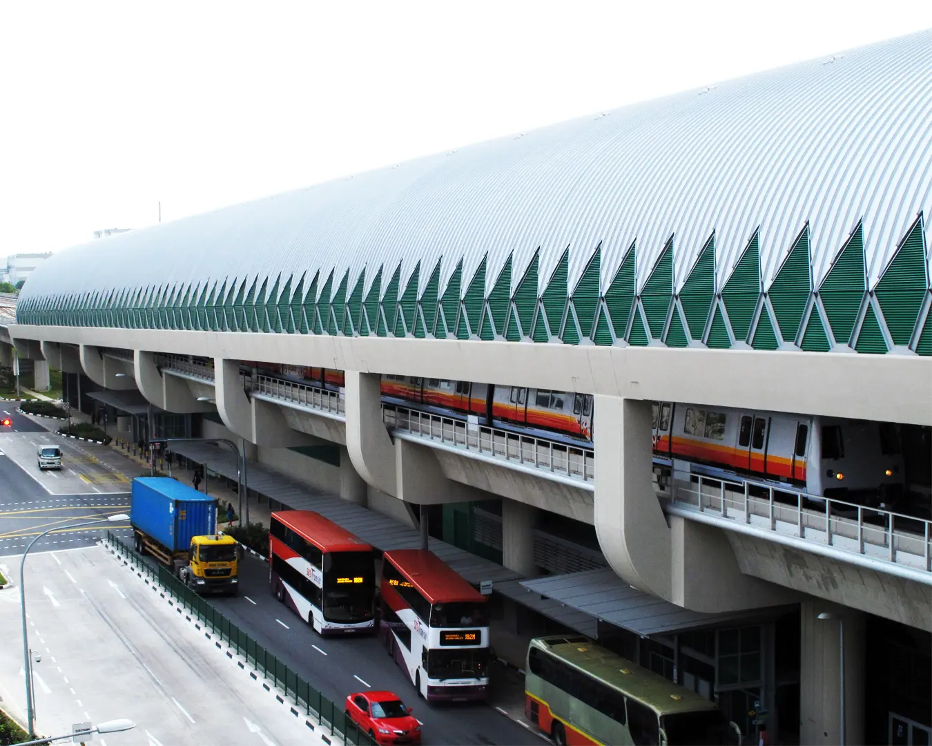 Elevated metro train station with a modern, ribbed roof. A colorful train travels above busy roads filled with buses and trucks, reflecting urban energy.