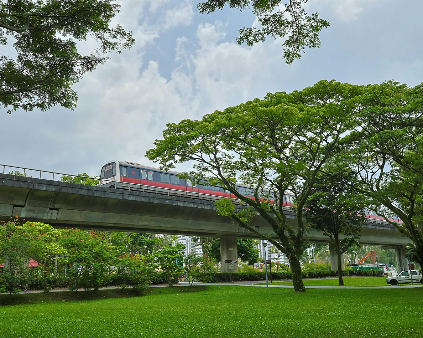 A red train travels on an elevated track through a lush, green park with tall trees. The sky is cloudy, creating a serene and peaceful atmosphere.