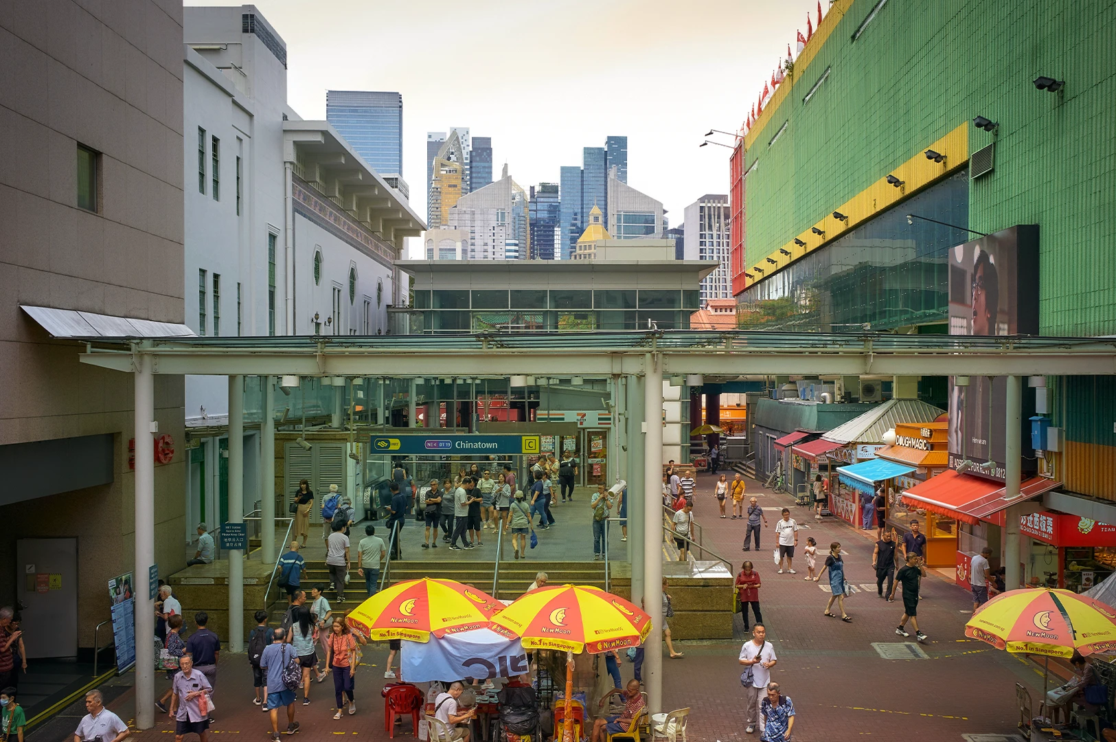 Busy pedestrian courtyard at Singapore's Chinatown MRT station exit, featuring colorful market stalls, people walking, and a backdrop of a modern city skyscrapers under a clear sky.
