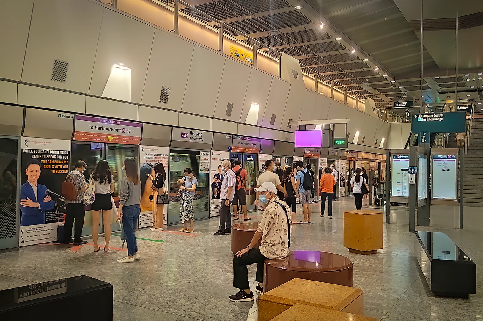 Modern underground platform of Hougang MRT station in Singapore, showing commuters waiting behind glass screen doors with directional signs for the North East Line toward HarbourFront.