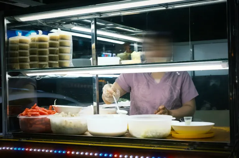 A candid photo of a street food vendor preparing an food at a night market stall, serving a plethora of fruits and cream under a bright display lighting.
