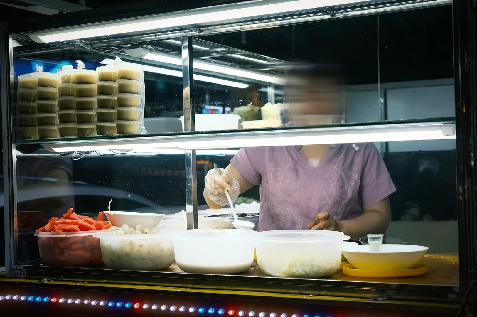 A candid photo of a street food vendor preparing an food at a night market stall, serving a plethora of fruits and cream under a bright display lighting.