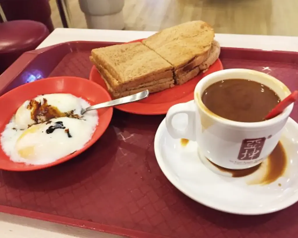 A breakfast tray with kaya toast on a red plate, soft-boiled eggs in another bowl, and a cup of coffee. A warm and inviting atmosphere.