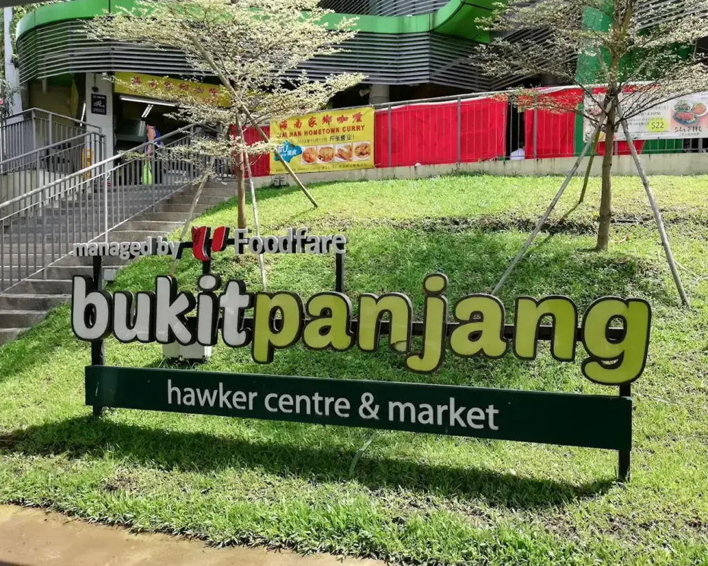 Sign for Bukit Panjang Hawker Centre & Market with trees and grass foreground. The building and entrance feature colorful banners behind.
