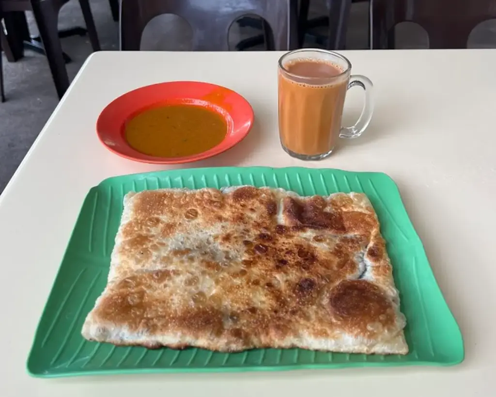 A crispy, square roti canai on a green plate beside a red dish of curry and a glass of milk tea on a white table. The setting is simple and inviting.