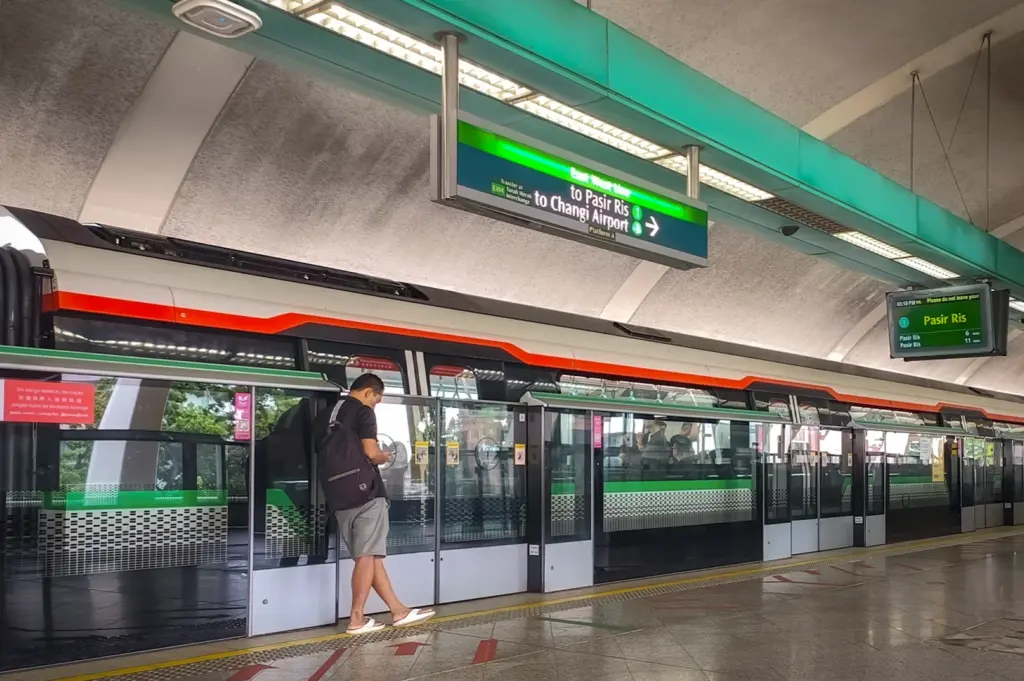 A vertical street-level shot of the Kembangan MRT station in Singapore, highlighting its iconic vaulted roof and elevated tracks, with a pedestrian standing near the train entrance.
