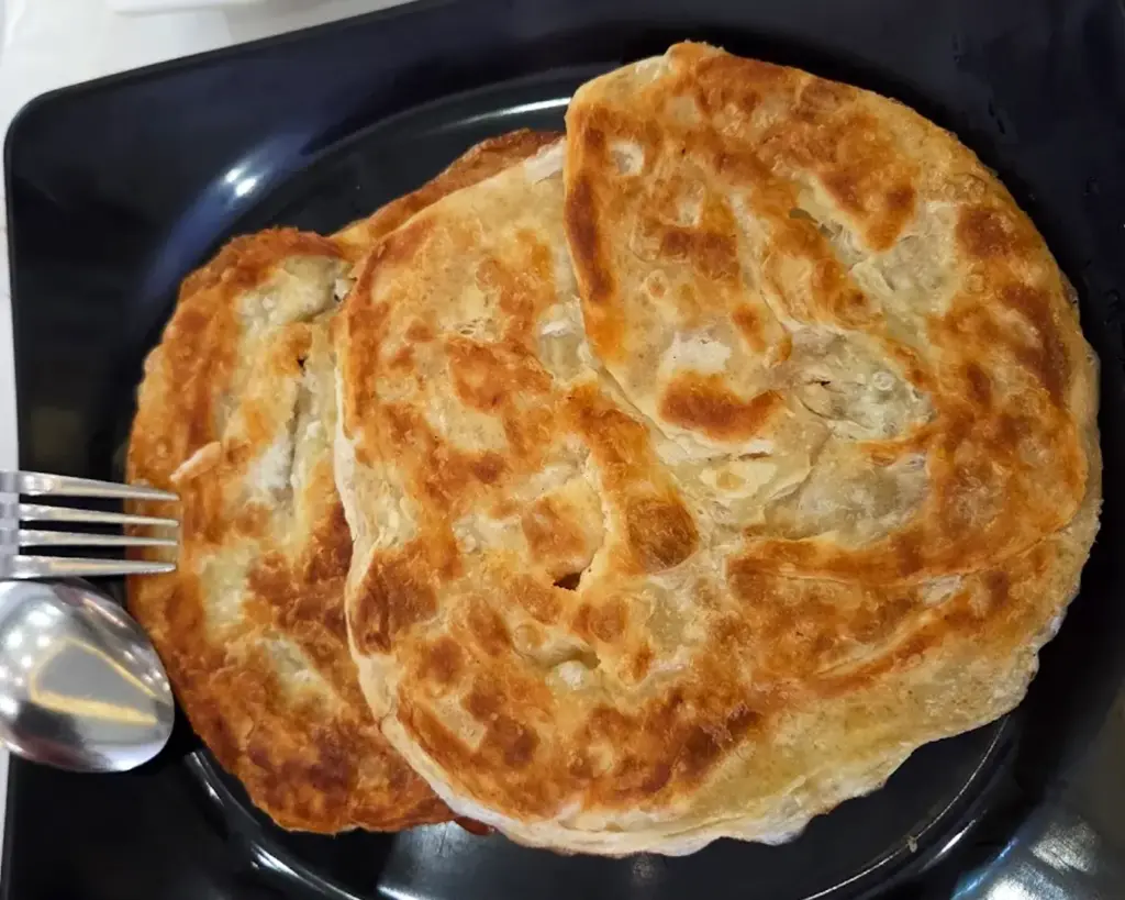 Two crispy, golden-brown flatbreads sit on a black plate, accompanied by a fork and a spoon. The tone is appetizing and warm.