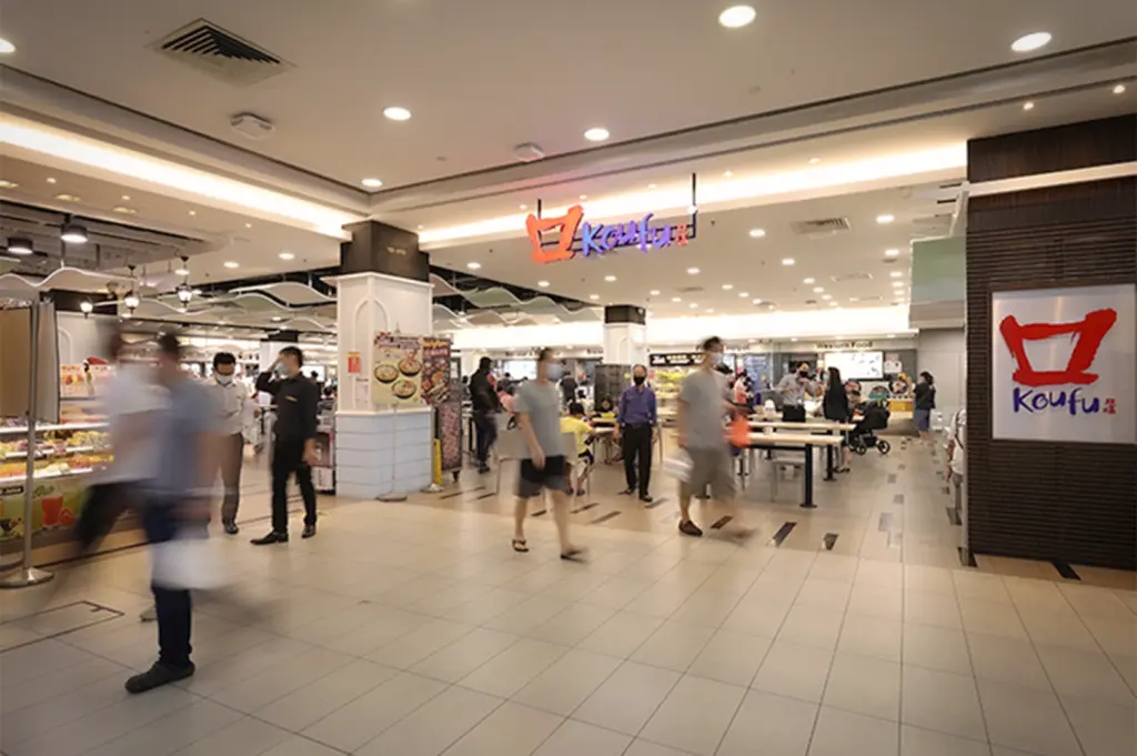 A bustling Koufu food court interior in a Singapore shopping mall, featuring blurred motion of people walking past food stalls and seating areas under bright modern lighting.