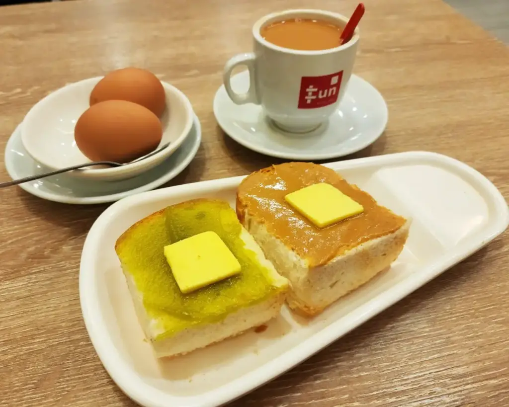 Toasted buns with kaya and peanut butter, each topped with butter, are on a white plate. Next to it are two soft-boiled eggs and a cup of tea.