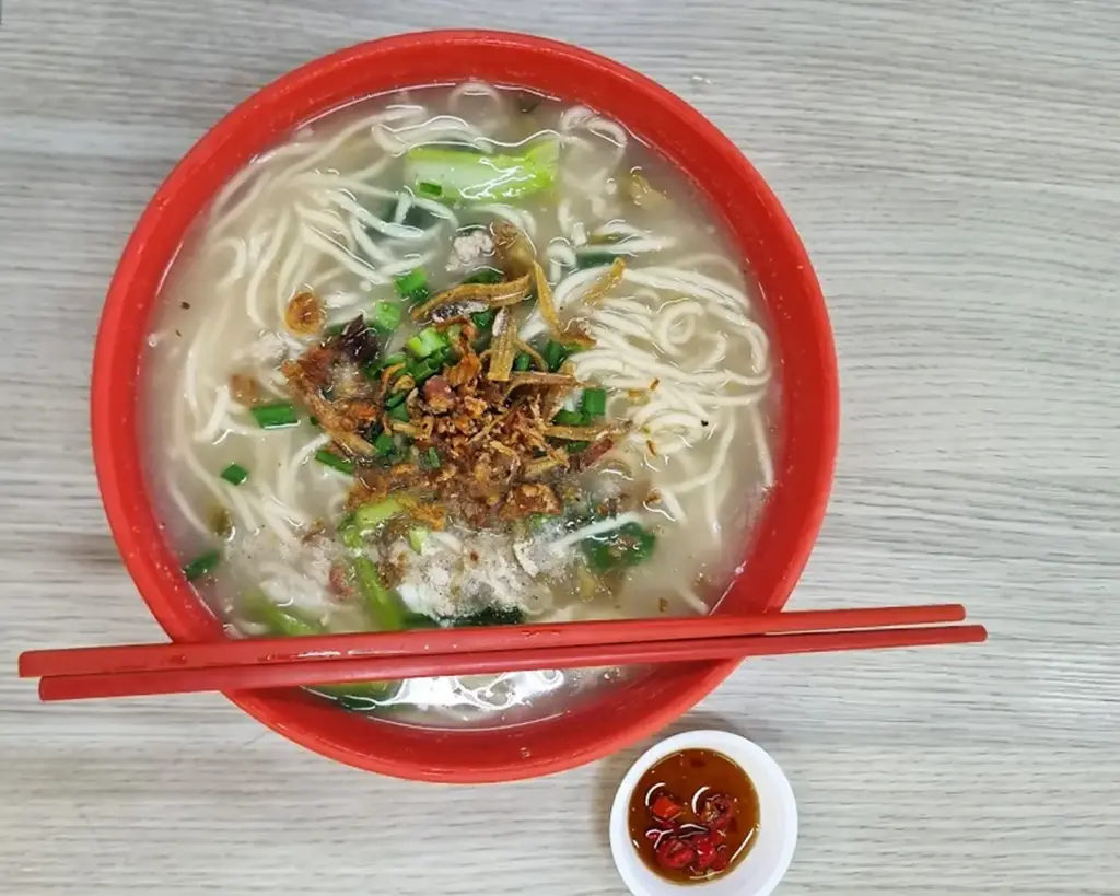 A red bowl of noodle soup with vegetables, crispy toppings, and red chopsticks placed on a light wooden table. A small dish of chili sauce is beside it.