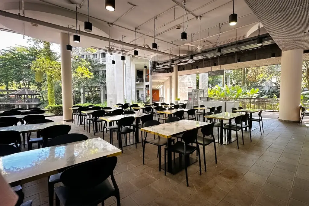 Modern open-air food court at Pioneer Hall, NTU Singapore, featuring marble-top tables, black chairs, and industrial pendant lighting overlooking a lush green landscape and campus architecture