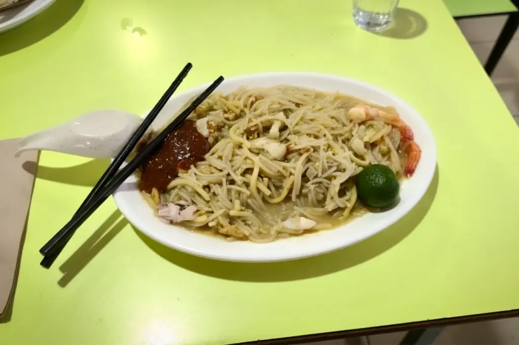 Authentic Singaporean Hokkien Prawn Mee served on a white plate, featuring stir-fried yellow noodles, rice vermicelli, fresh prawns, squid, and a side of spicy sambal chili and calamansi.
