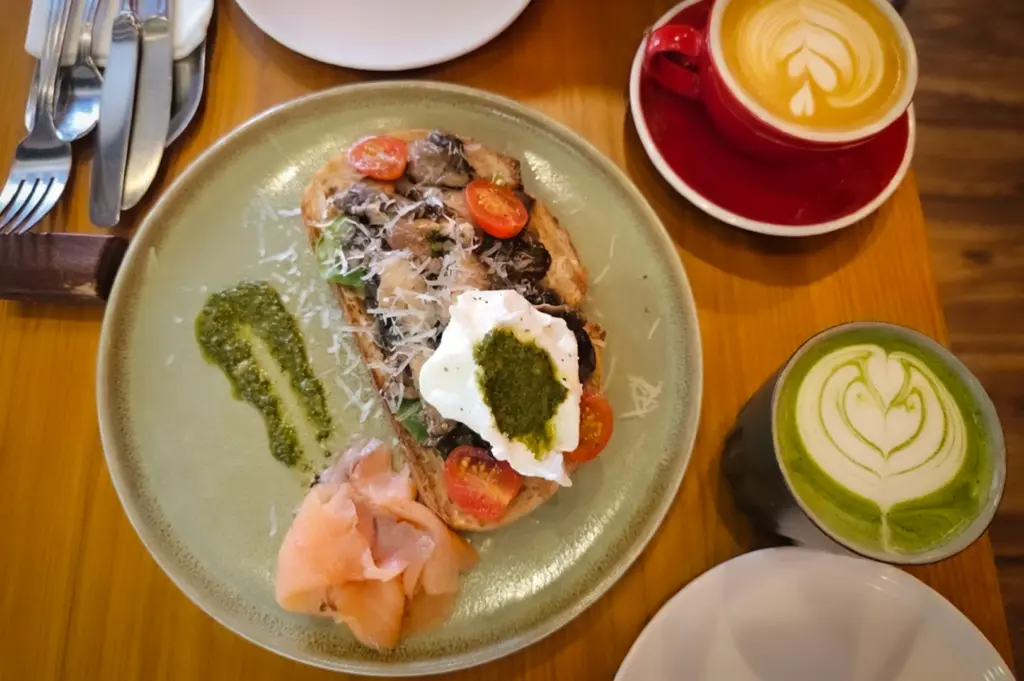 A delicious brunch spread from Refuel Cafe, featuring mushroom sourdough toast topped with a poached egg, pesto, and cherry tomatoes, served with a side of smoked salmon, accompanied by a hot latte and a matcha green tea latte with latte art.