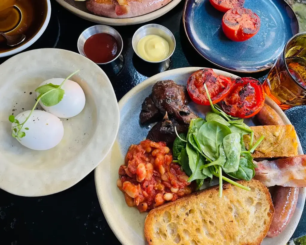A hearty breakfast spread with poached eggs, grilled tomatoes, sausages, bacon, baked beans, spinach, toast, and sauce sides on a black table.