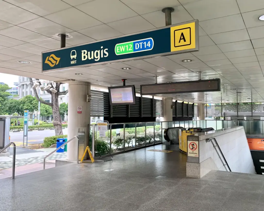 Entrance to Bugis MRT station in Singapore, featuring directional signs, an escalator, and nearby greenery. The scene feels modern and organized.