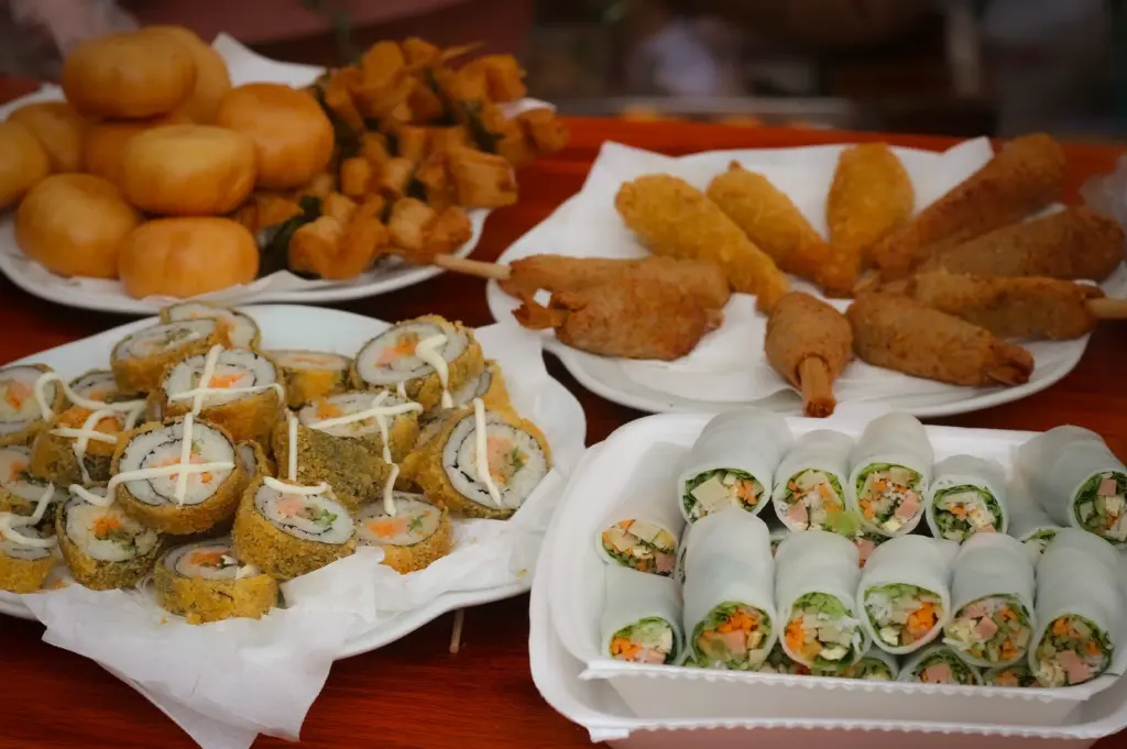 A variety of Asian street food including fresh Vietnamese spring rolls, deep-fried sushi rolls with mayo, golden fried mantou buns, and crispy skewers served on a wooden table.