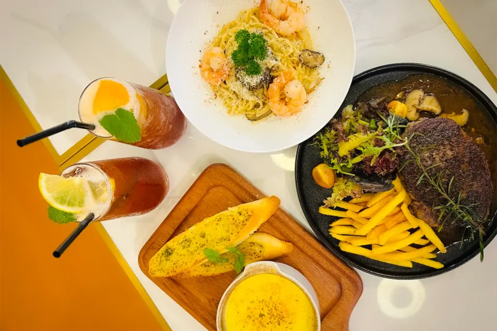 A vibrant cafe meal spread featuring prawn aglio olio, a juicy steak served with fries and fresh garden salad, and golden garlic bread with a side of creamy soup, paired with refreshing iced fruit teas at C Cafe in Joo Chiat.