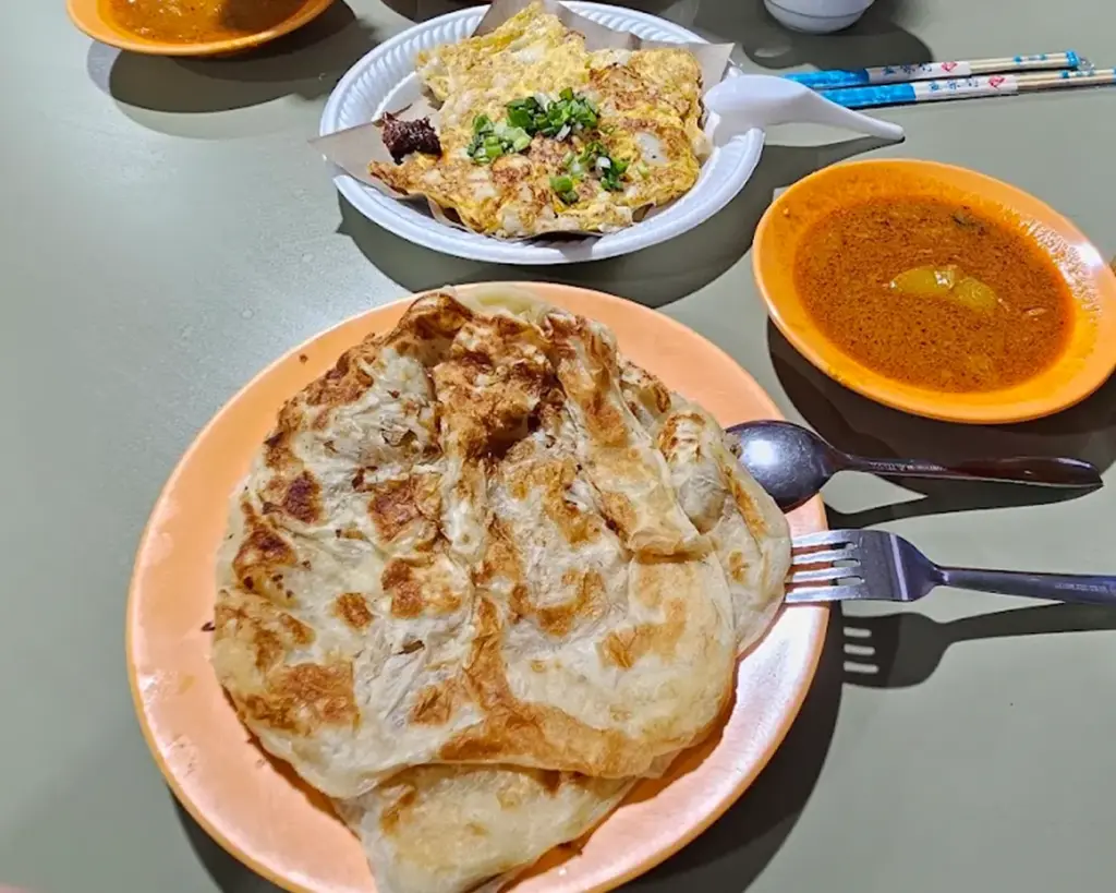 A plate of golden, flaky roti and another with an omelette topped with onions and greens are shown. A small bowl of orange curry is on the side.