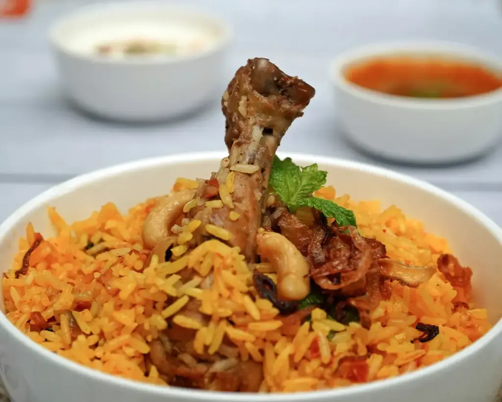 A bowl of vibrant orange biryani with a chicken drumstick, topped with fried onions, cashews, and mint. Two bowls of curry and yogurt are in the background.