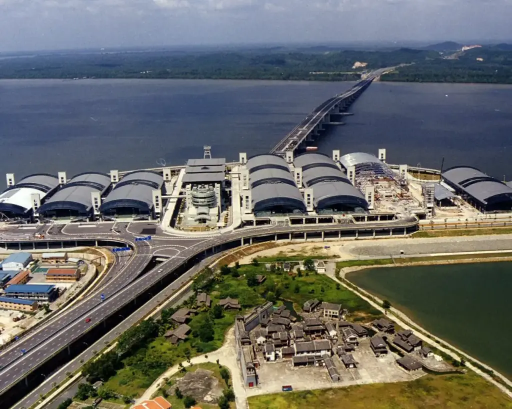 Aerial view of a large transport terminal with multiple domed structures, connecting to a long bridge over water. Roads and greenery surround the area.