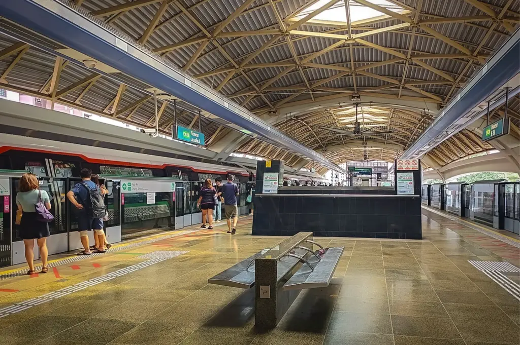 Elevated platform of Pioneer MRT station in Singapore featuring a waiting train, commuters, and a distinct industrial arched roof with metal trusses and ceiling fans.