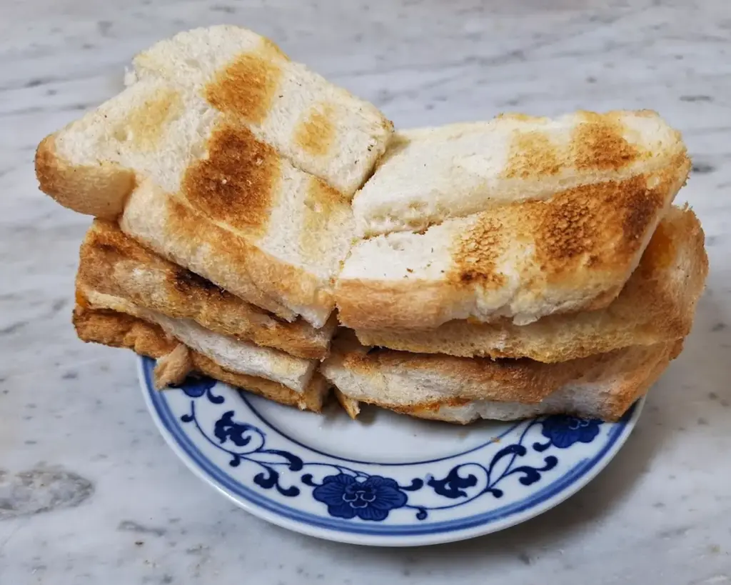 A small stack of grilled white bread slices on a blue and white floral plate, placed on a marble surface. The bread is golden brown with grill marks.