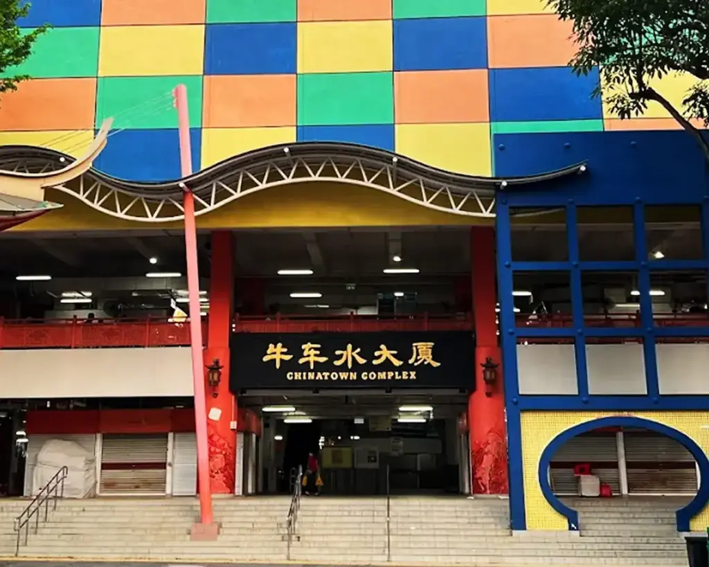 Colorful facade of Chinatown Complex with red, blue, green, and yellow tiles. Arched entrance features signage in English and Chinese. Steps lead up.