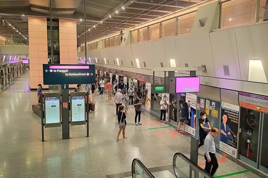 Wide-angle view of the modern Hougang MRT station platform in Singapore, featuring commuters waiting for North East Line trains, directional signage toward Punggol and HarbourFront, and glass safety screen doors.