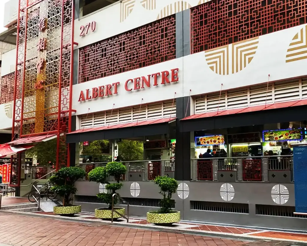 Exterior of Albert Centre with red Chinese lattice details and a large sign. Food stalls visible inside. Decorative potted plants line the walkway.
