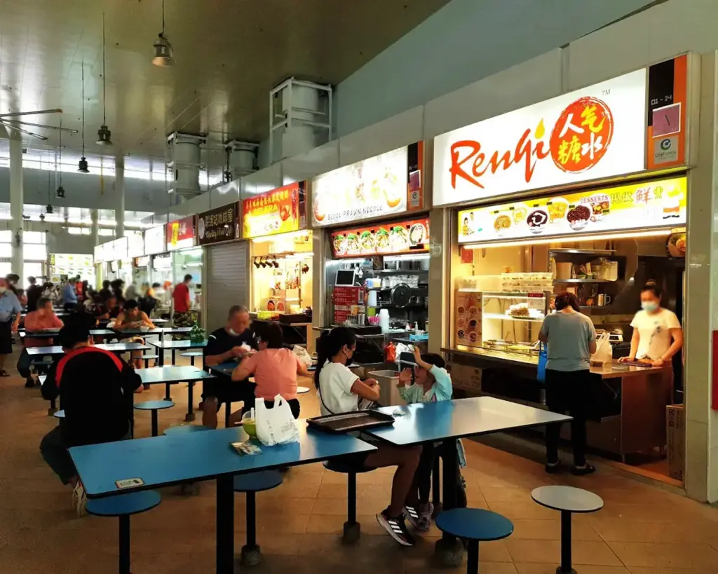 A bustling food court with people sitting at blue tables, eating and chatting. Brightly lit food stalls in the background display various Asian cuisines.